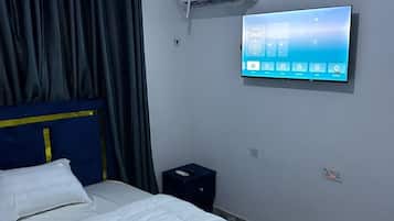 Habitación de hotel con cama, televisor de pantalla plana en la pared, equipo de aire acondicionado y mesita de noche con lámpara.
