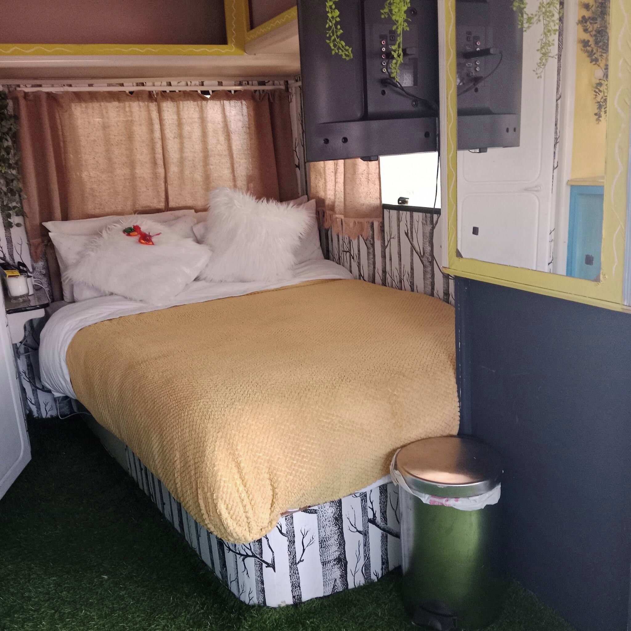 Uma cama com um cobertor branco e bege, uma pequena lixeira e uma janela com cortinas.
