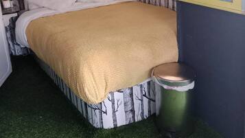 Uma cama com um cobertor branco e bege, uma pequena lixeira e uma janela com cortinas.