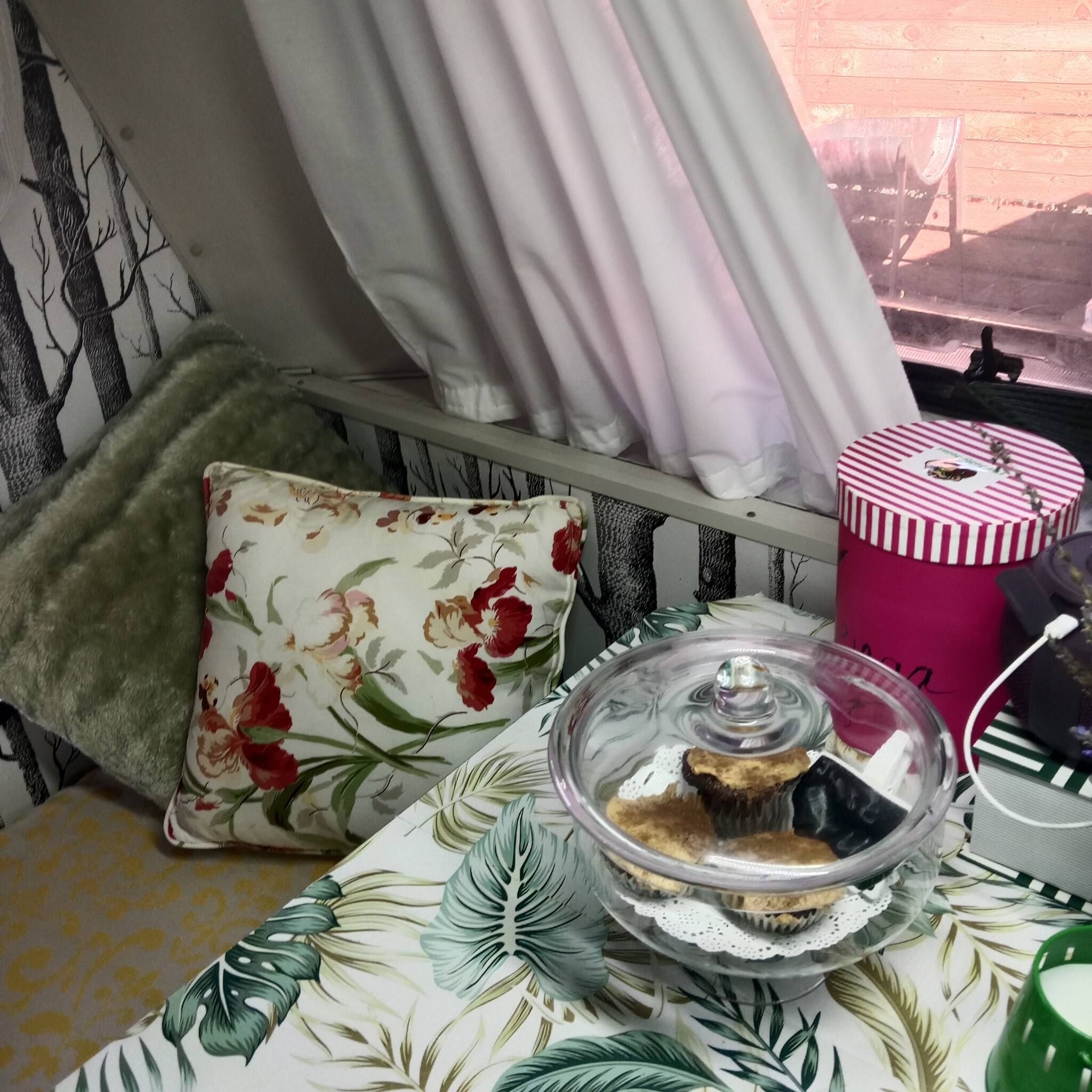 Een kamer met een bed, een raam met gordijnen, een kussen, een tafel met een glazen vaas en een gestreepte container.