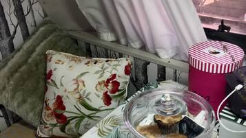 Een kamer met een bed, een raam met gordijnen, een kussen, een tafel met een glazen vaas en een gestreepte container.