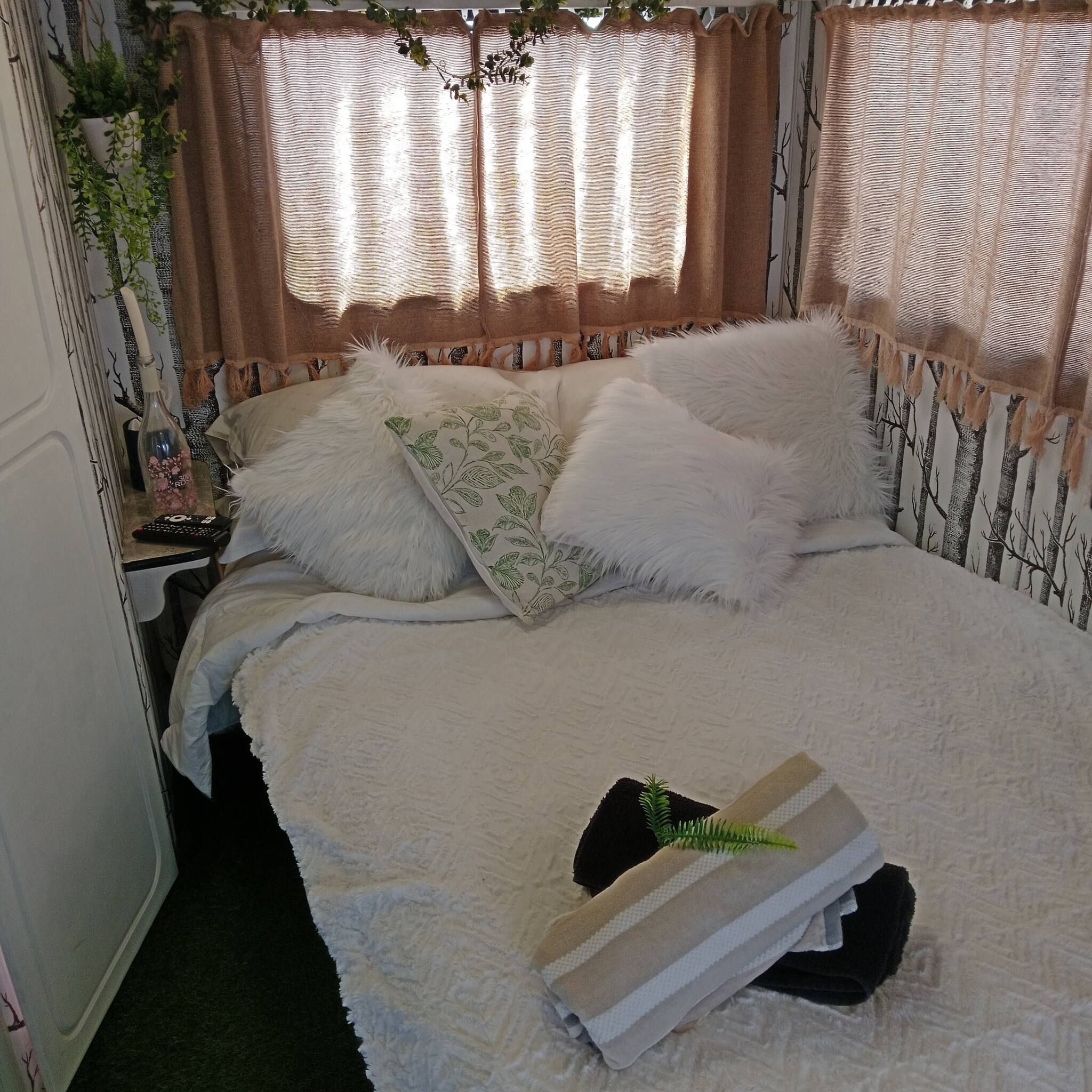 Een bed met witte beddengoed en twee kussens, een nachtkastje met een fles en afstandsbediening, en een raam met gordijnen.