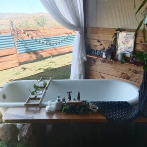 Casa de banho com banheira, uma prateleira de madeira com plantas e vista para um deck de madeira e colinas.