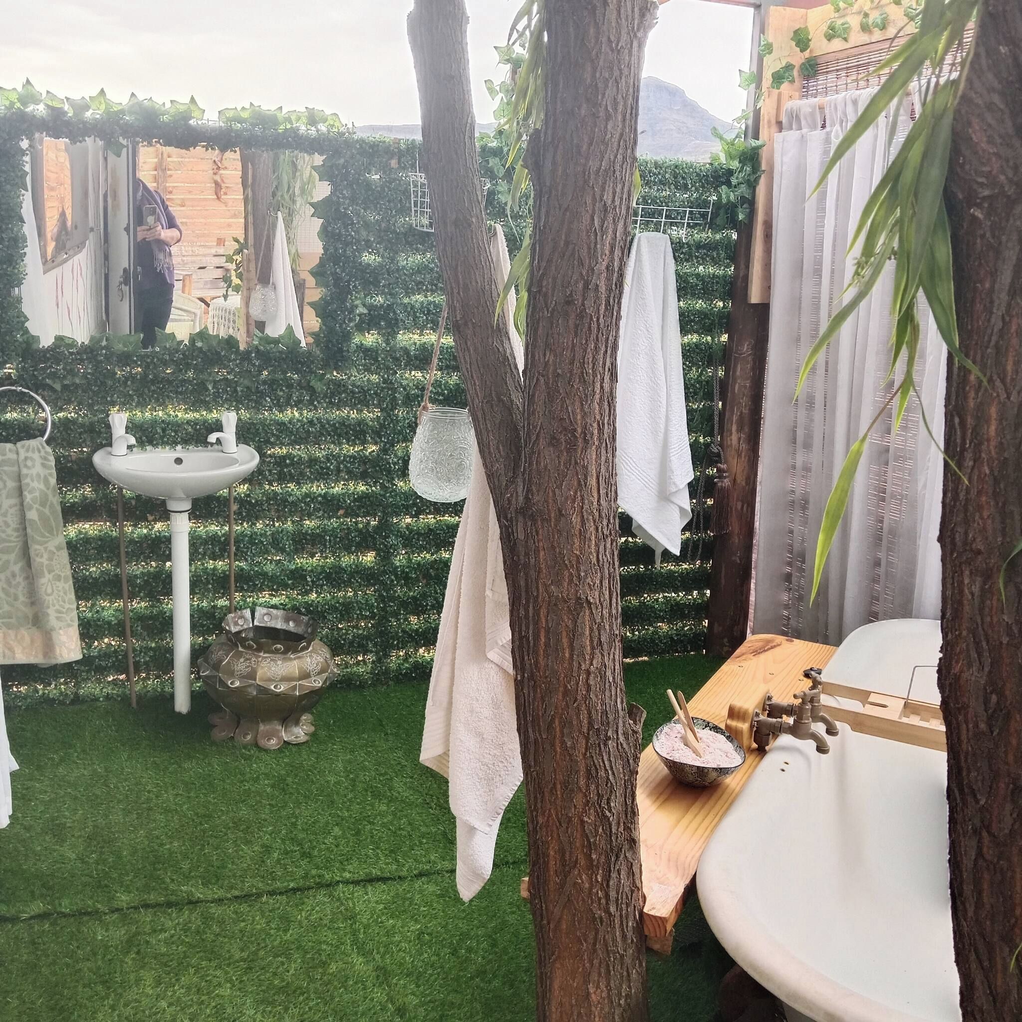 Un baño con lavamanos, espejo e inodoro. Se ve un árbol en primer plano y un seto al fondo.