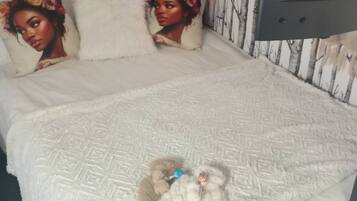 Ein Bett mit einer weißen Decke, zwei Kissen, einem Teddybären und einem Fenster mit Vorhängen.