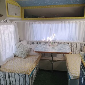 A cozy RV interior with a dining table, cushioned seating, and a window with curtains. - Romantic Clarens Getaway - Glamped Caravan With Braai & Fairy Bath (Clarens)