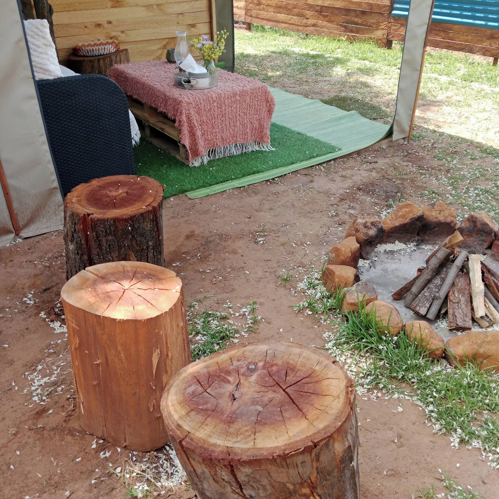 Un espacio de estar al aire libre rústico con un banco de madera, una mesa pequeña con un jarrón de flores y un brasero rodeado de leña y rocas.