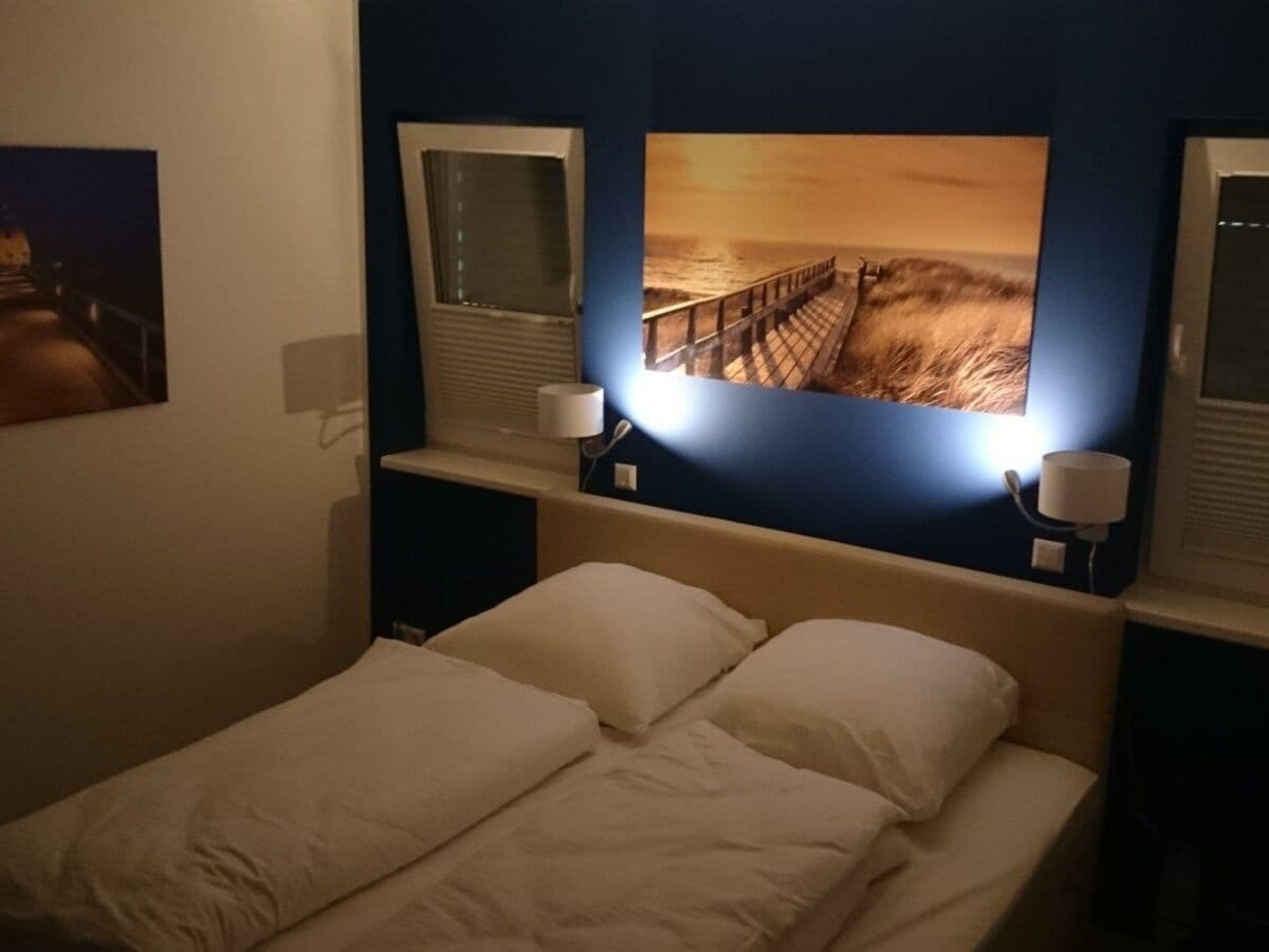 Habitación de hotel con una cama, dos lámparas de noche y un cuadro grande de una puesta de sol sobre el océano.