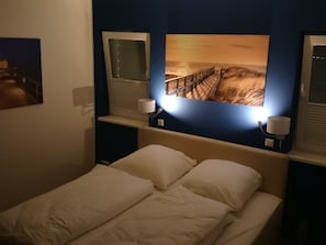 A hotel room with a bed, two bedside lamps, and a large picture of a sunset over the ocean. - "captain Dornfelder" (Trent)