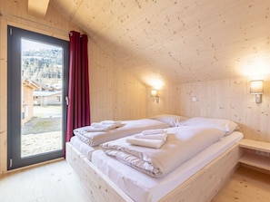 Um quarto de madeira com cama, janela com cortinas vermelhas e vista para edifícios lá fora.