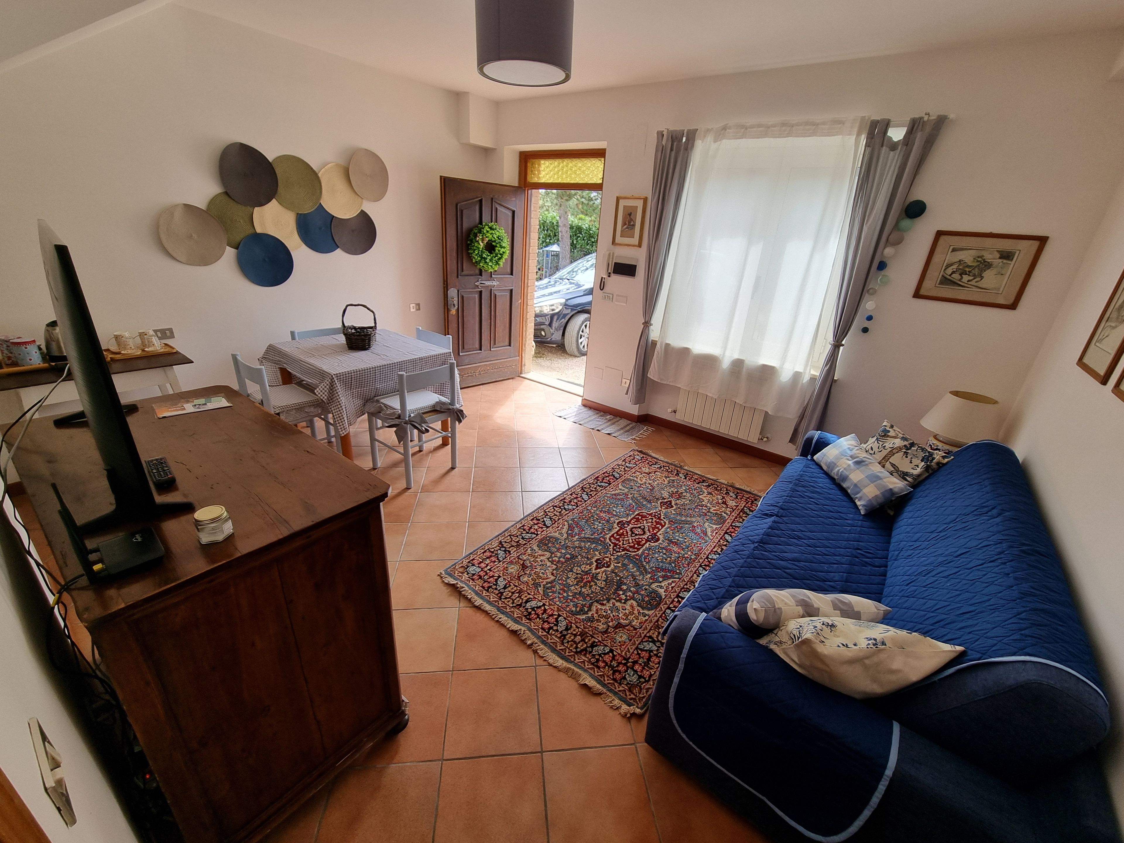 A living room with a blue sofa, a wooden TV stand, a dining table with chairs, a patterned rug, and a door leading outside.