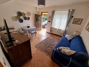 Una sala de estar con un sofá azul, un mueble de televisión de madera, una mesa de comedor con sillas, una alfombra estampada y una puerta que da al exterior.