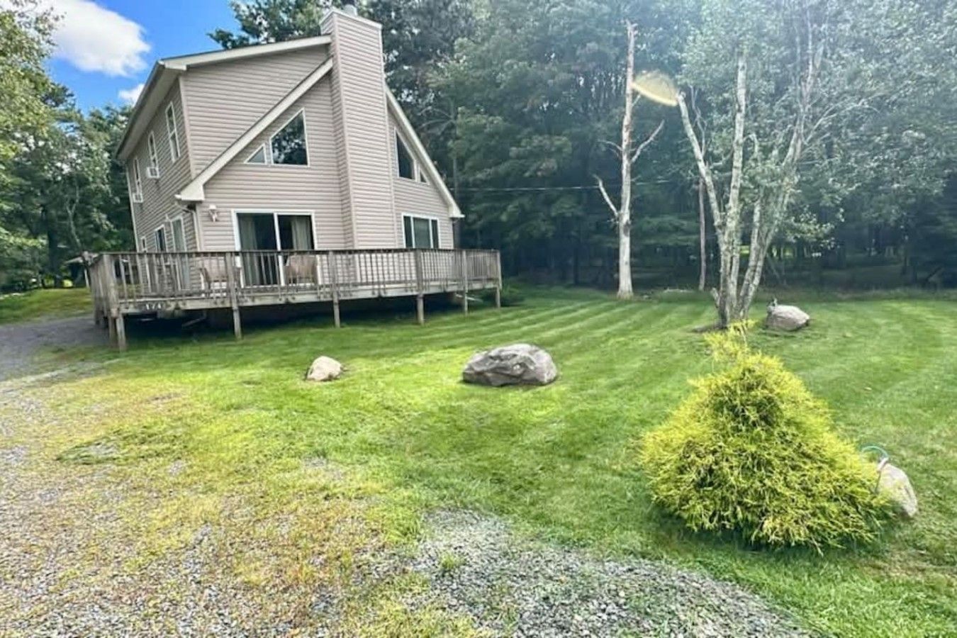 A two-story house with a deck, surrounded by a well-maintained lawn with rocks and a small bush.
