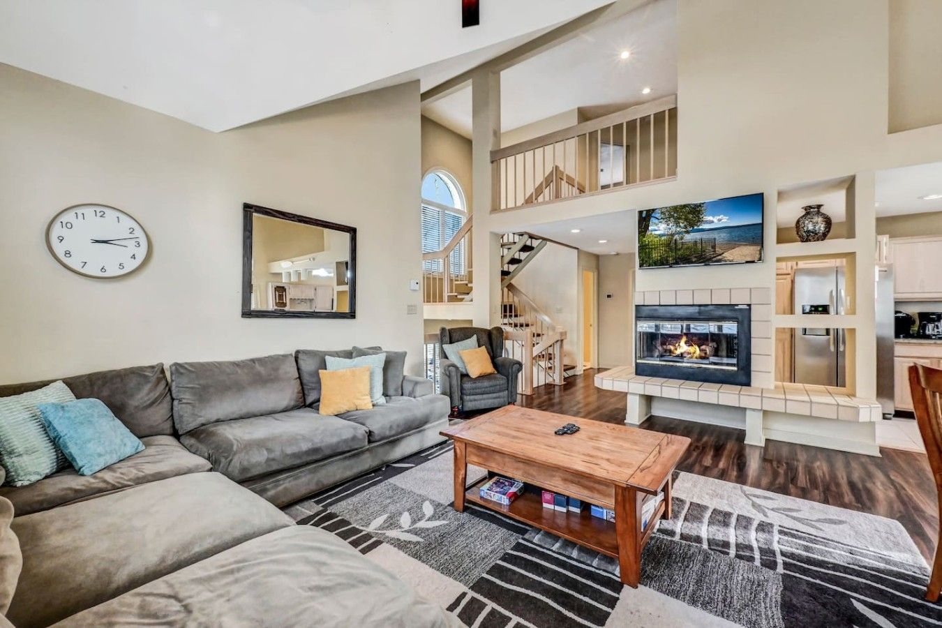 A spacious living room with a grey sofa, a coffee table, and a fireplace. A staircase leads to an upper level.