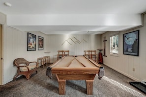 Una mesa de billar en una habitación con paredes blancas y suelo alfombrado. Hay dos sillones y una mesita con una botella.