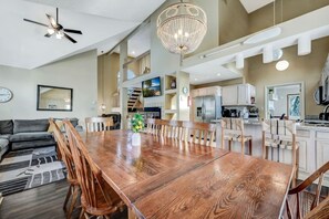Een ruime woonkamer met een houten eettafel, zachte zithoek, een kroonluchter en een moderne keuken op de achtergrond.
