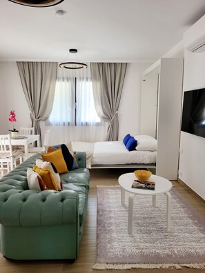Un saló modern amb un sofà verd, un llit blanc, una taula blanca petita i un televisor gran a la paret.