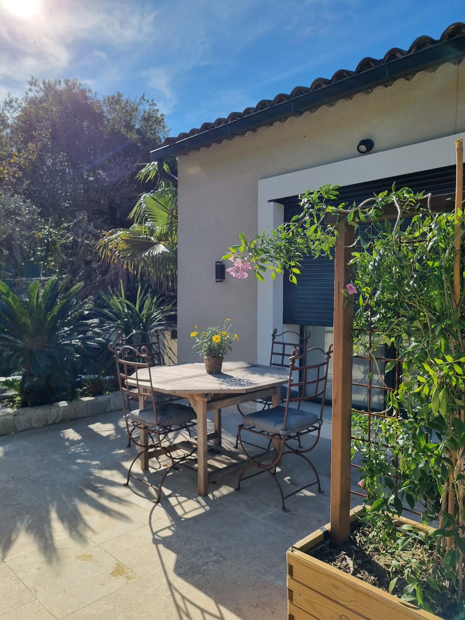 Eine Terrassengaststätte mit Tisch und Stühlen, umgeben von Pflanzen und einem Gebäude mit Ziegeldach.