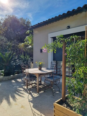 Eine Terrassengaststätte mit Tisch und Stühlen, umgeben von Pflanzen und einem Gebäude mit Ziegeldach.