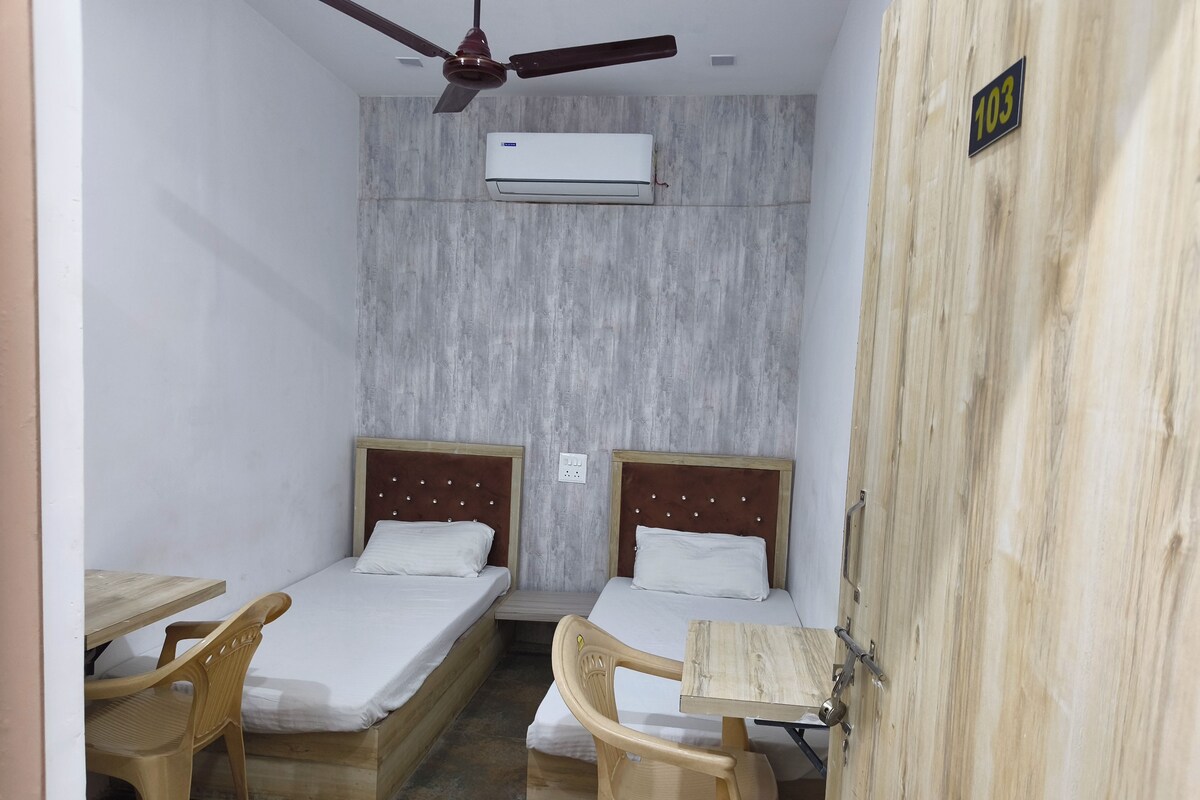 A room with two beds, a ceiling fan, an air conditioning unit, and a wooden door with a number sign.
