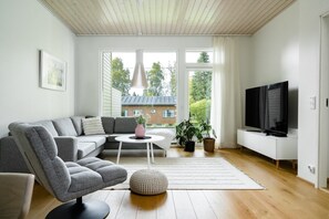 A modern living room with a grey sectional sofa, a white coffee table, a television, and a large window with a view of a house and trees.