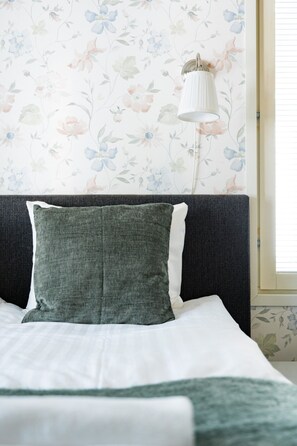 A bed with a green pillow and white bedding, a dark headboard, and a floral wallpaper pattern.