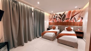 Quarto de hotel com duas camas, uma janela grande com cortinas, parede com padrão de folhas e televisão de tela plana fixada na parede.