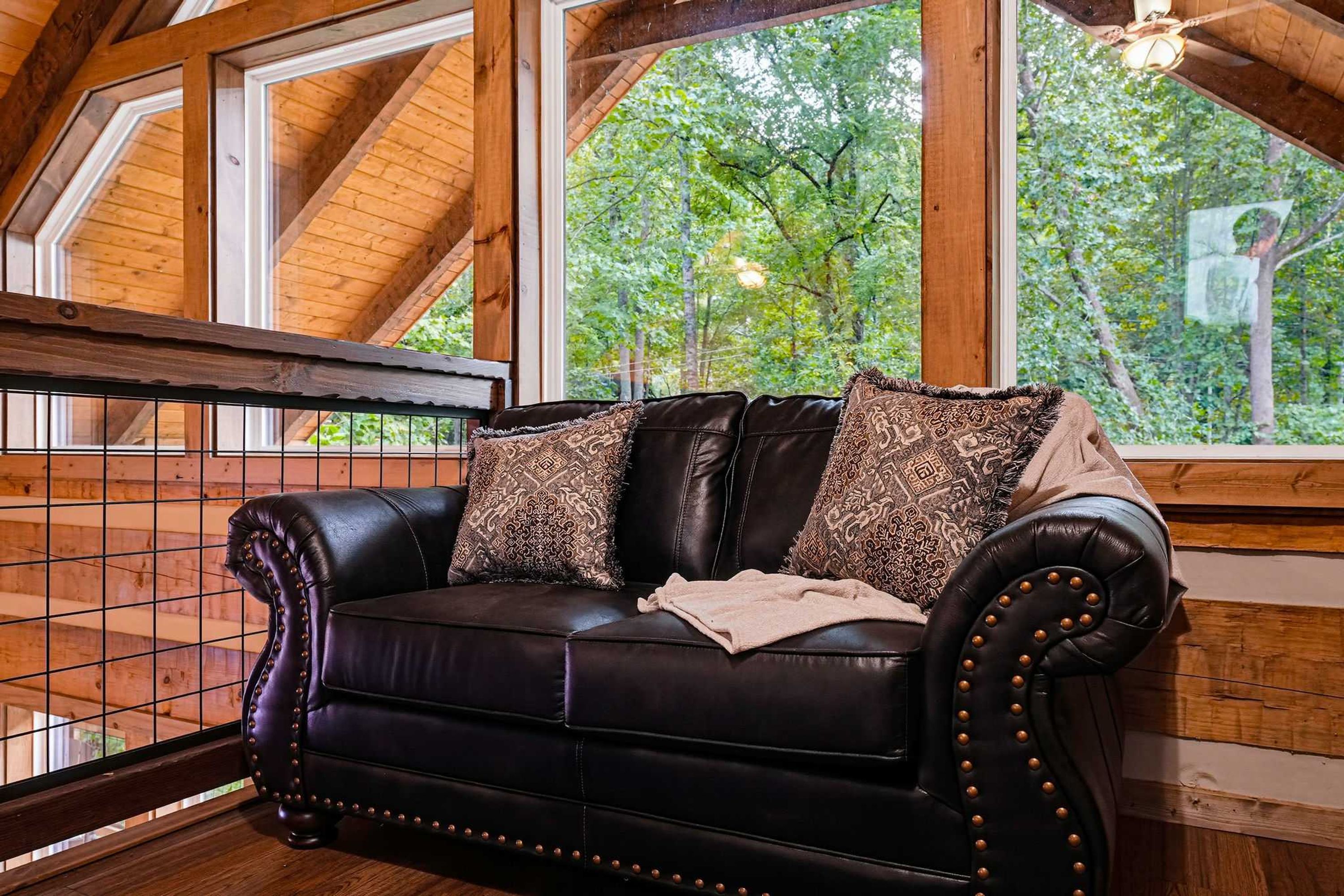 A black leather sofa with decorative pillows and a throw blanket, positioned in a room with wooden beams and large windows overlooking a forest.
