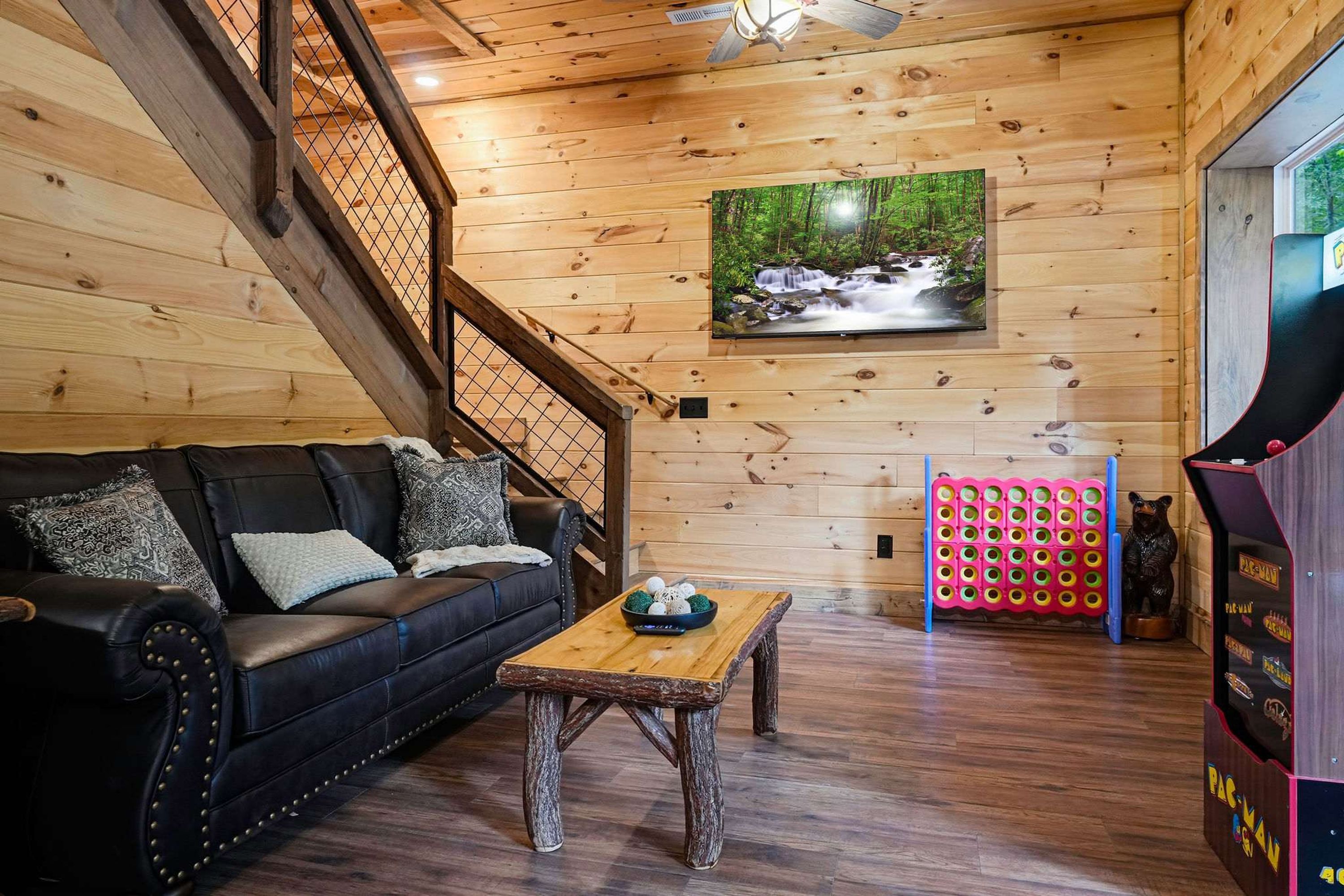 A wooden interior with a black leather sofa, a wooden coffee table, a wall-mounted TV, and a Pac-Man arcade machine.