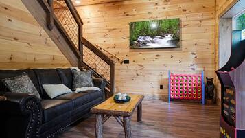 A wooden interior with a black leather sofa, a wooden coffee table, a wall-mounted TV, and a Pac-Man arcade machine.