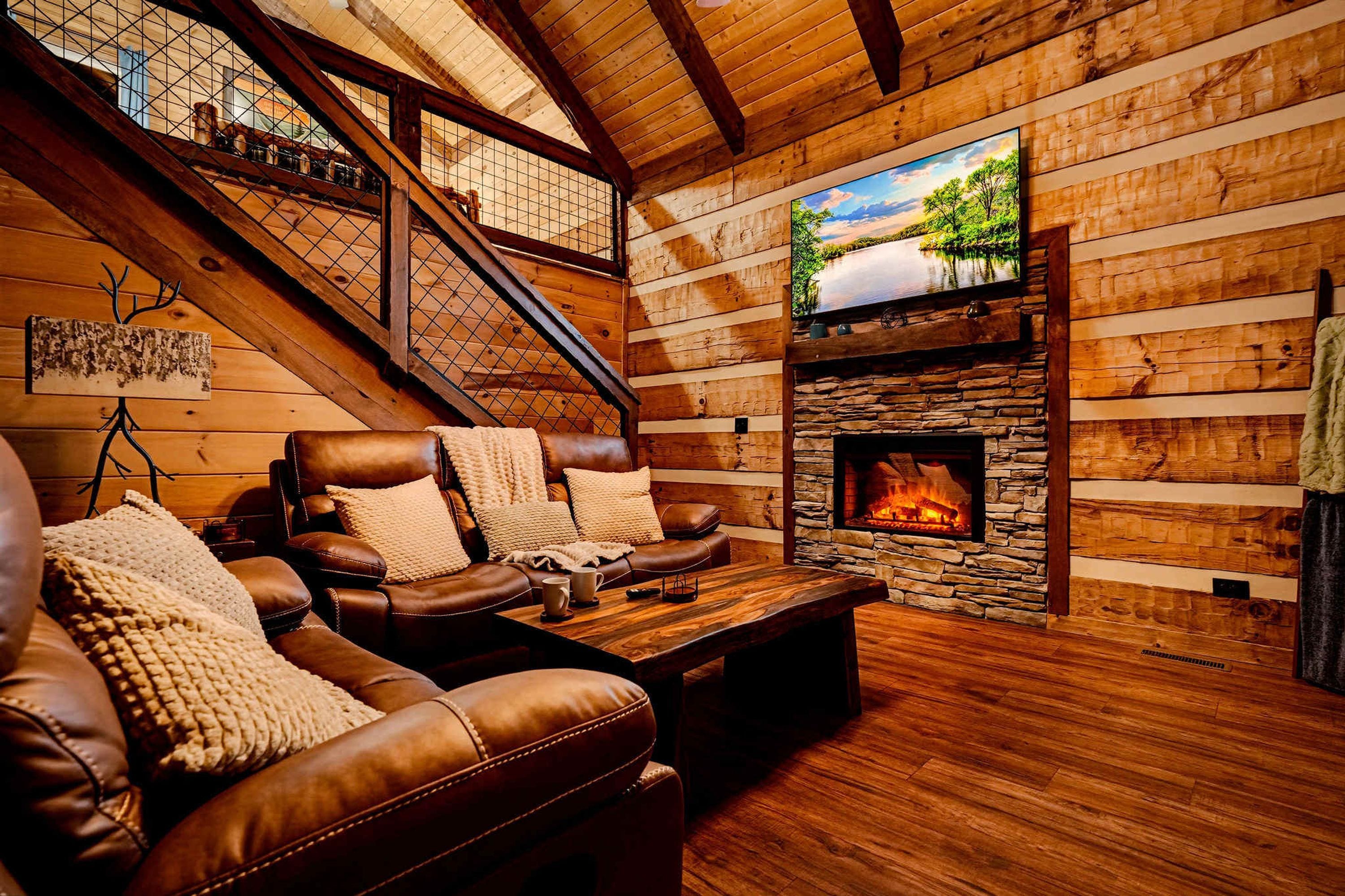 A cozy cabin interior with a stone fireplace, leather seating, and a wooden staircase.