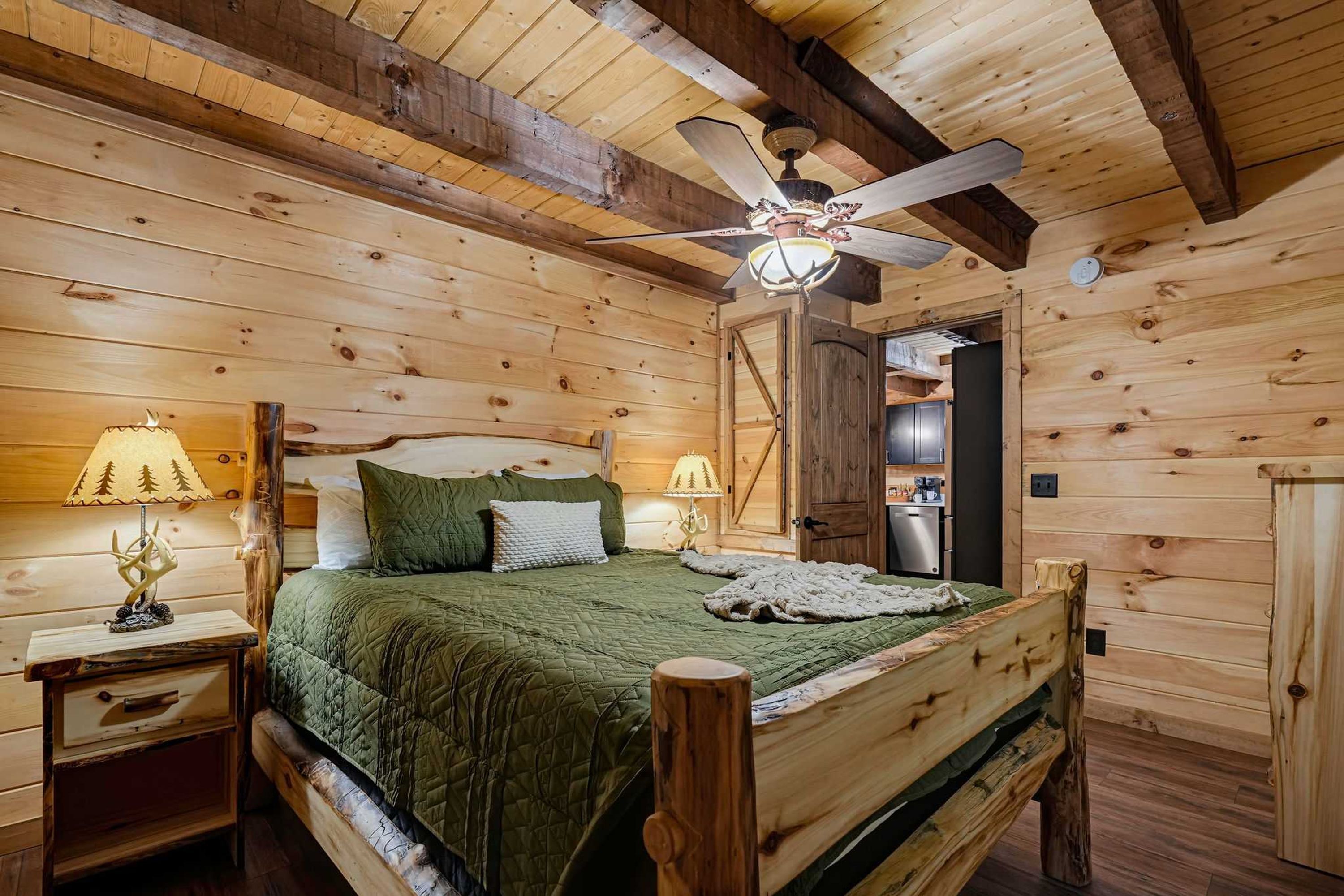 Un dormitorio de cabaña de madera con una cama, dos lámparas de noche, un ventilador de techo y una puerta que da al baño.
