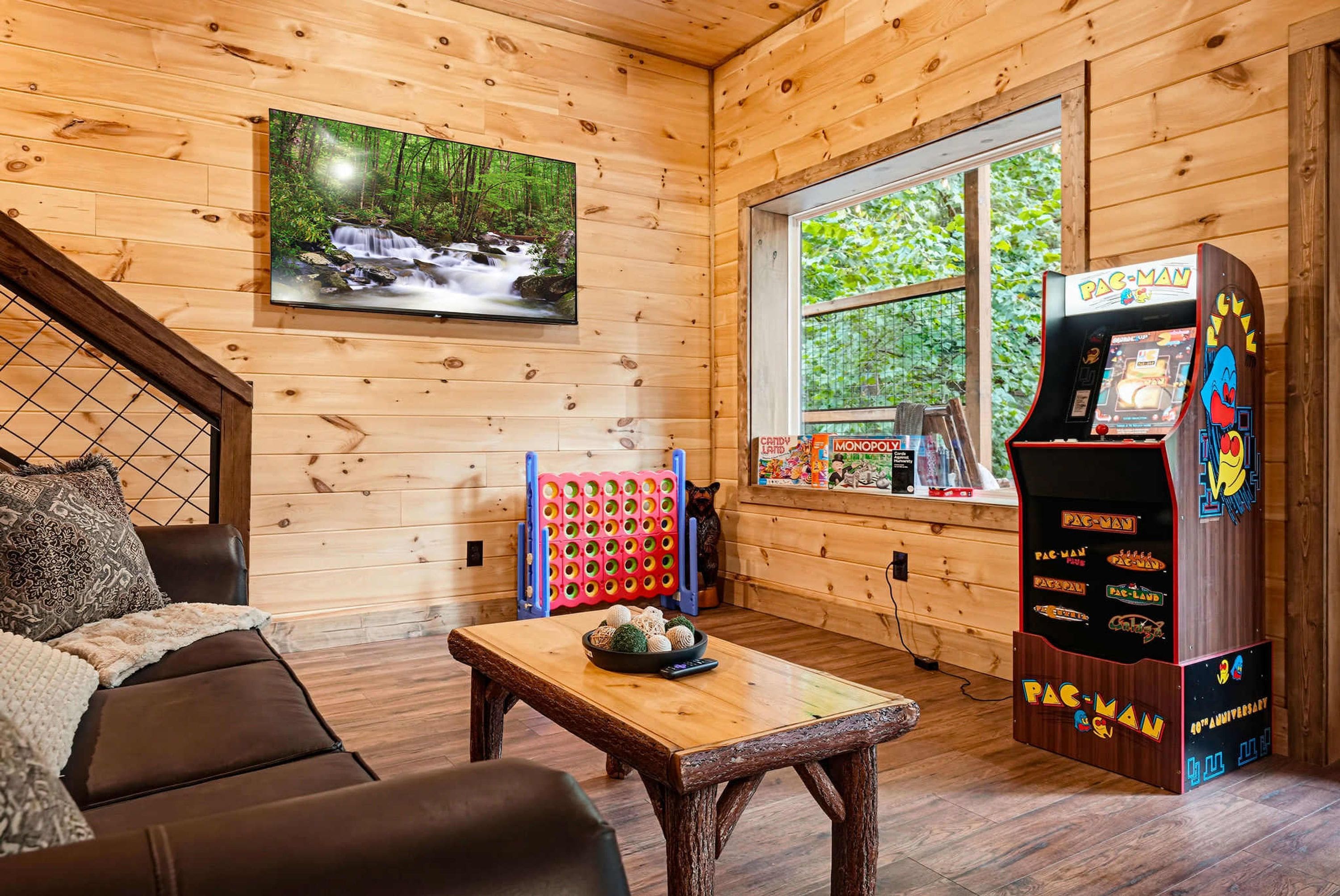 Habitació acollidora amb interior de fusta, televisió penjada a la paret, una màquina recreativa de Pac-Man i una finestra amb vistes a la verdor.