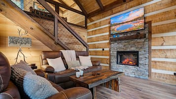 A cozy log cabin interior with a stone fireplace, leather sofa, and a large TV mounted on the wall.