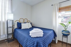 A neatly made bed with a blue quilt, a bedside table with a remote control, and a potted plant on a small table beside the window. - Great Walkable Location! Tucson Studio Stay (Tucson)