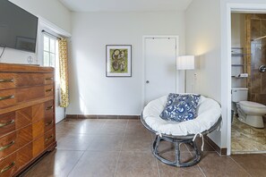 A room with a wooden dresser, a framed picture on the wall, a white door, a round chair with a cushion, and a bathroom with a toilet and shelves. - Great Walkable Location! Tucson Studio Stay (Tucson)