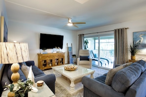 A living room with a sofa, coffee table, and a television. There is a ceiling fan, a cabinet with decorative items, and a view of the outside through a glass door. - Access to Pool & Sanibel Beach: Resort-style Condo (Sanibel)
