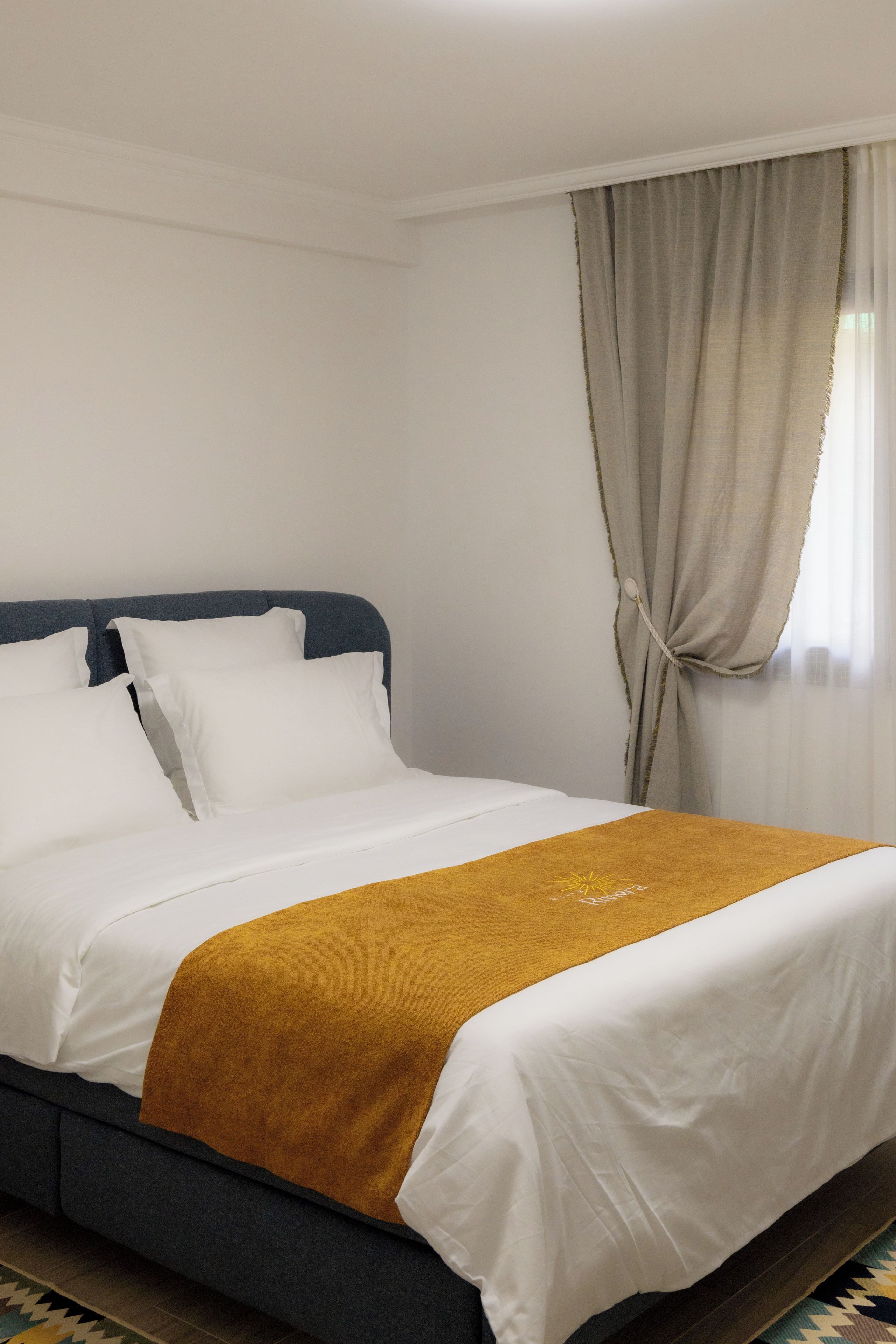 Ein Hotelzimmer mit Bett, weißer Bettwäsche, einer senfgelben Tagesdecke und einem Fenster mit Vorhängen.