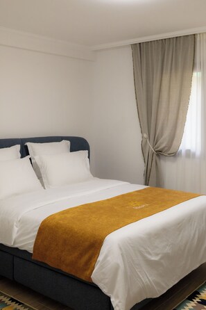 Ein Hotelzimmer mit Bett, weißer Bettwäsche, einer senfgelben Tagesdecke und einem Fenster mit Vorhängen.