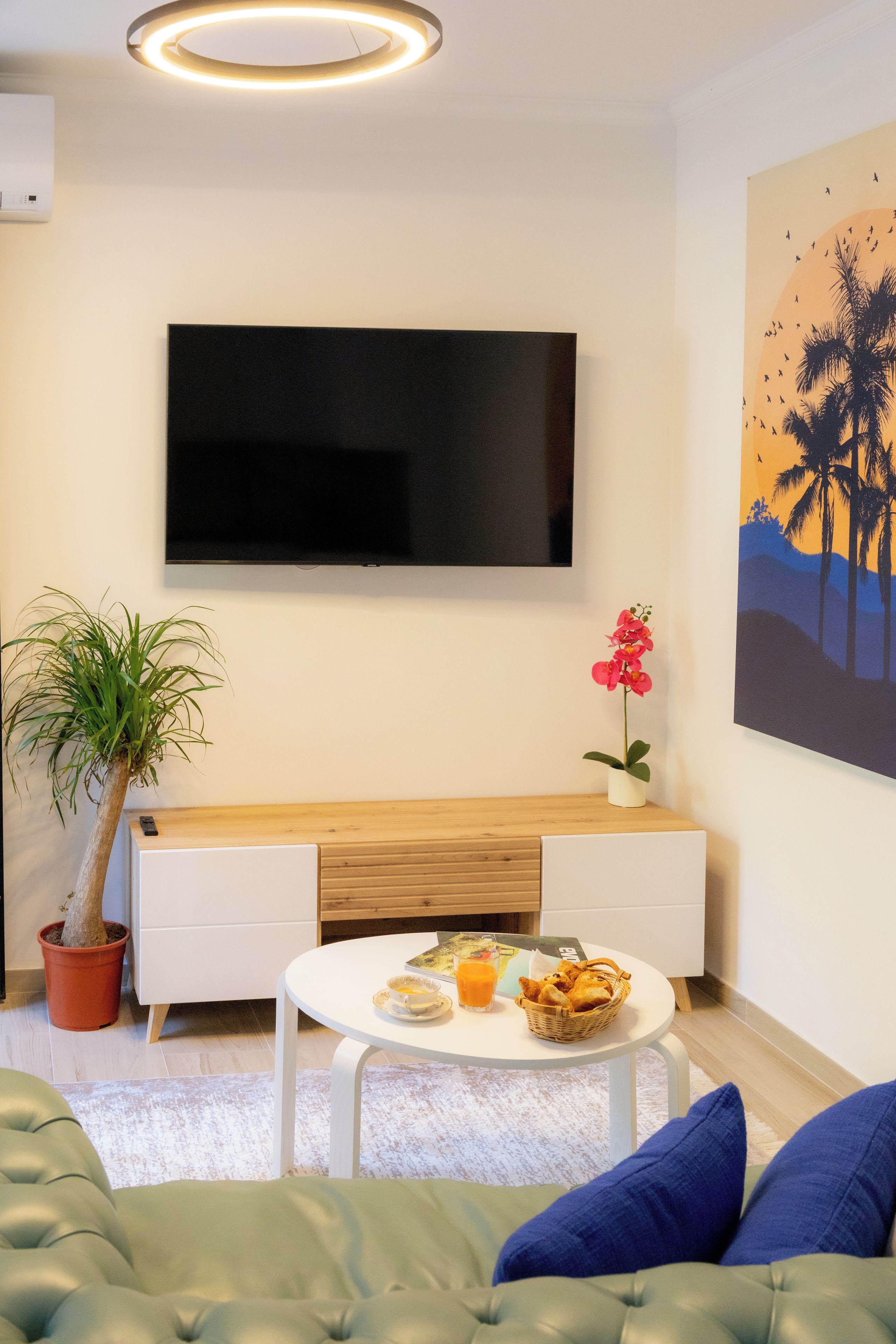 Ein modernes Wohnzimmer mit einem an der Wand befestigten Flachbildfernseher, einer Holzbank, einer Topfpflanze, einem kleinen Tisch mit einem Brotkorb und einem Palmen-Gemälde.