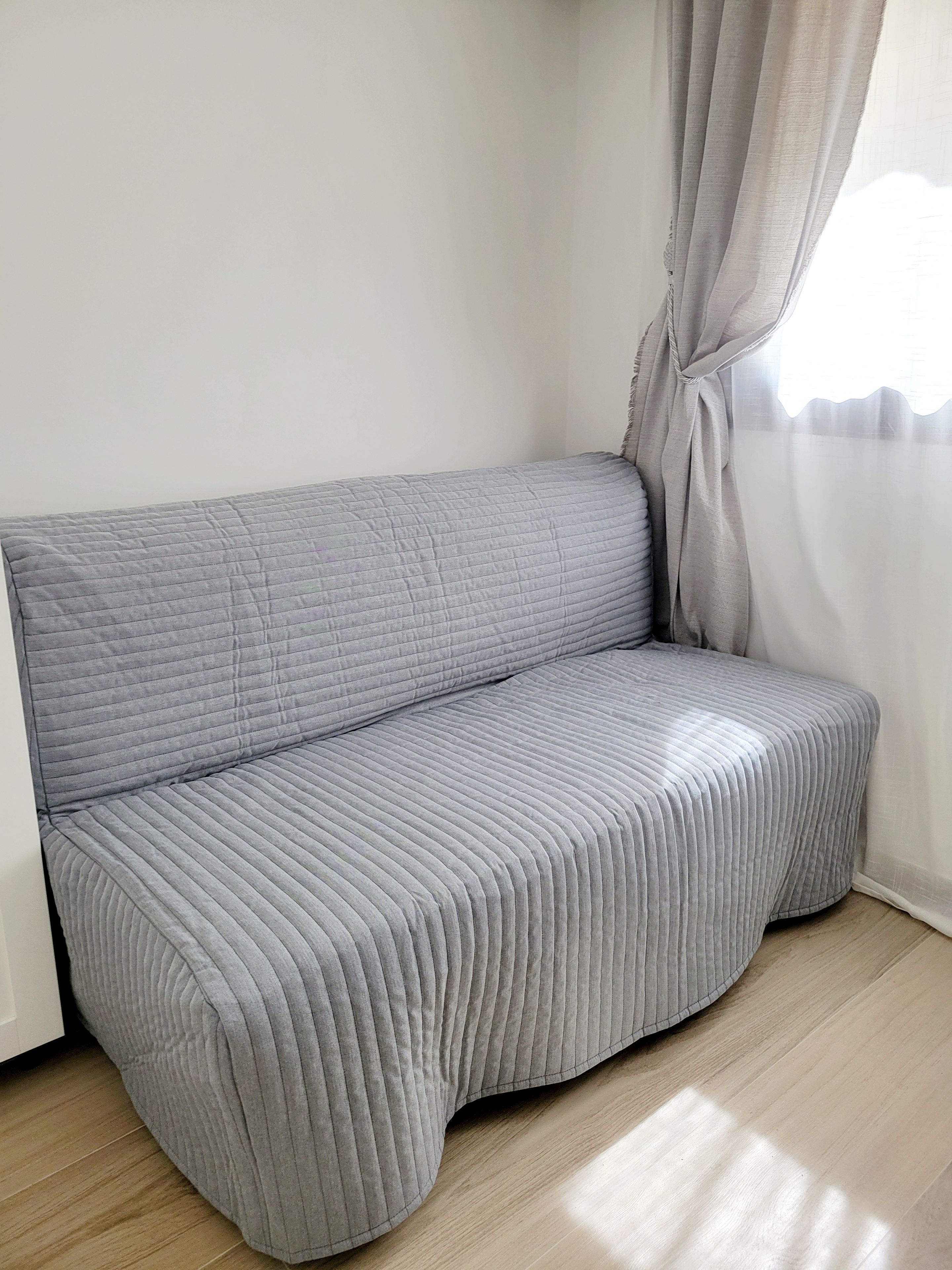 Ein graues, gepolstertes Sofa mit gerippter Struktur, platziert an einer weißen Wand mit Fenster und durchsichtigen Vorhängen.