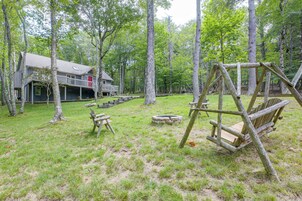 A rustic wooden house with a deck, surrounded by a forested area with a swing set and a fire pit.