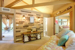 Quarto aconchegante com parede de madeira, cama com roupa de cama estampada, mesa com abajur e vista para um banheiro.