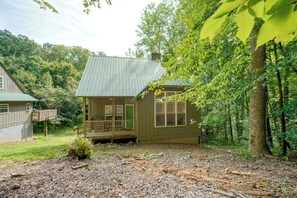 Une petite cabane dotée d’un porche, entourée d’arbres et bordée d’une zone boisée.