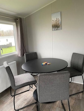 Ein Zimmer mit einem runden Tisch, vier grauen Stühlen, einem Heizkörper, einem Fenster mit Vorhängen und einem Wandbild.