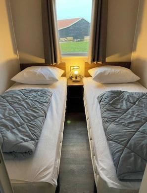 Ein kleines Zimmer mit zwei Betten, einem Nachttisch mit Lampe und einem Fenster mit Blick auf ein Gebäude und einen Hügel.