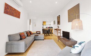 Ein modernes Wohnzimmer mit einem grauen Sofa, einem Couchtisch und einem Fernseher. Es gibt einen Essbereich mit Tisch und Stühlen. Die Wände sind weiß und der Boden ist aus Holz.