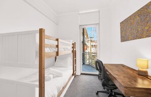 Ein Zimmer mit einem Etagenbett, einem Schreibtisch mit Stuhl, einer Lampe und einem großen Fenster mit Blick auf Gebäude.