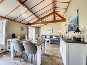 Ein geräumiger Raum mit hoher Decke, Holzbalken, einer Essecke mit Stühlen und Tisch, einem Sofa und einem Gemälde an der Wand.