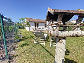 Ein eingezäunter Gartenbereich mit einem weißen Gebäude im Hintergrund, einer hölzernen Konstruktion und einem Liegestuhl.